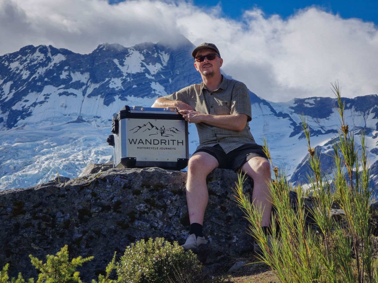 Man in cap, sunglasses, check shirt and shorts on a boulder, arm on a Wandrith-branded aluminium case, glacier ice and snowy peaks behind