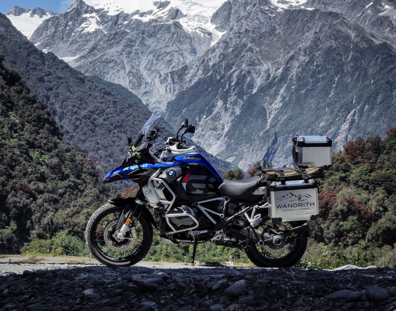 BMW R1250GS Adventure with Wandrith luggage on river stones, below rainforest-covered slopes and grey rock walls topped with snow and ice