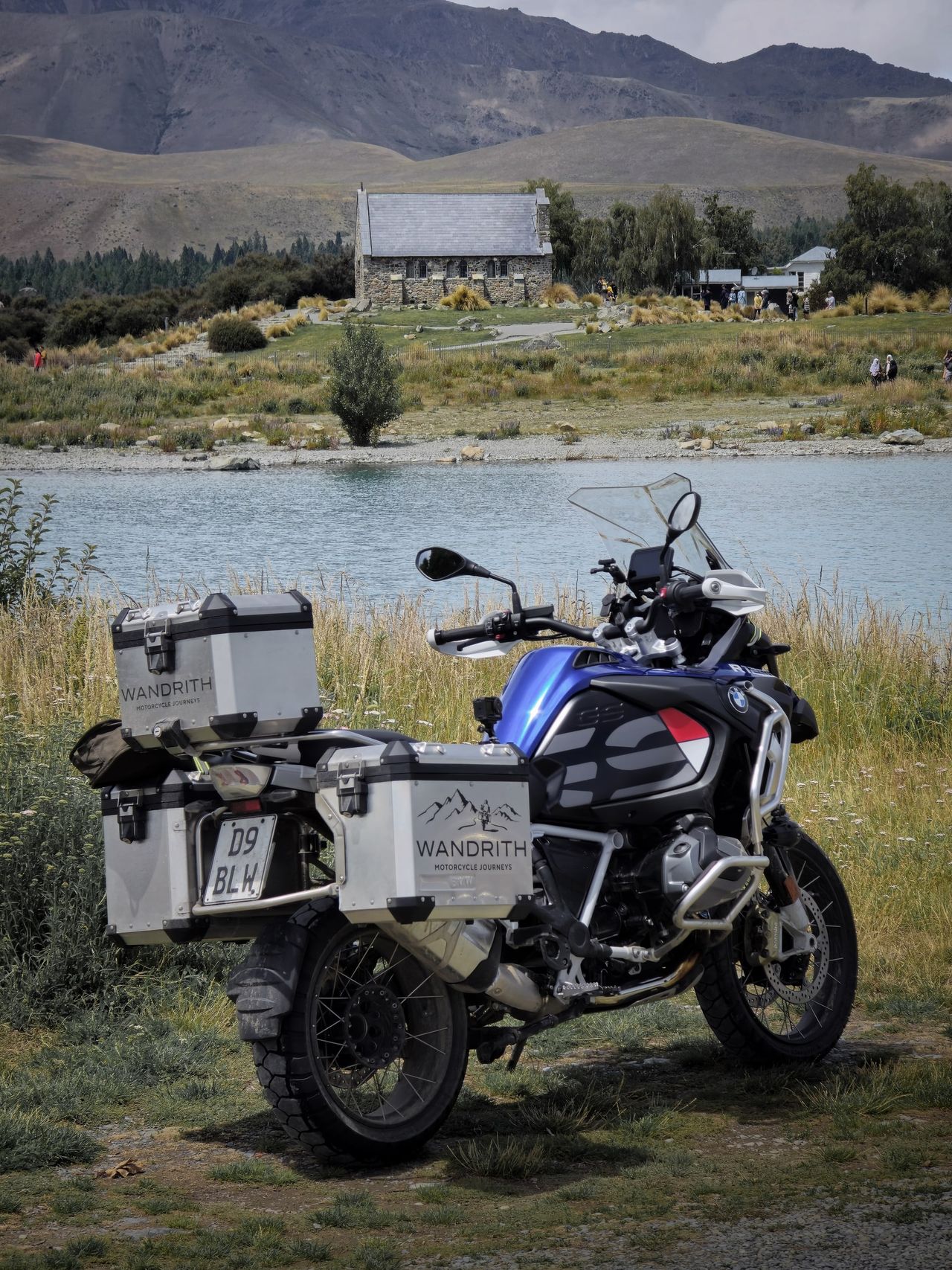 BMW R 1250 GS Adventure with Wandrith panniers parked in long grass by Lake Tekapo, a small stone church across the water