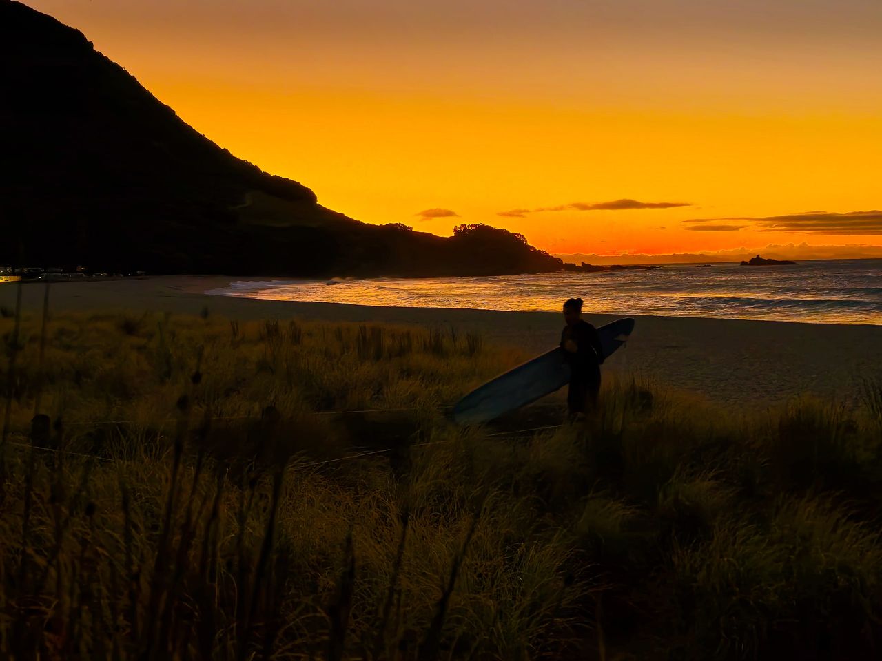 Silhouette of a surfer carrying a longboard through dune grass at sunset, beside the dark slope of Mauao and a golden sky