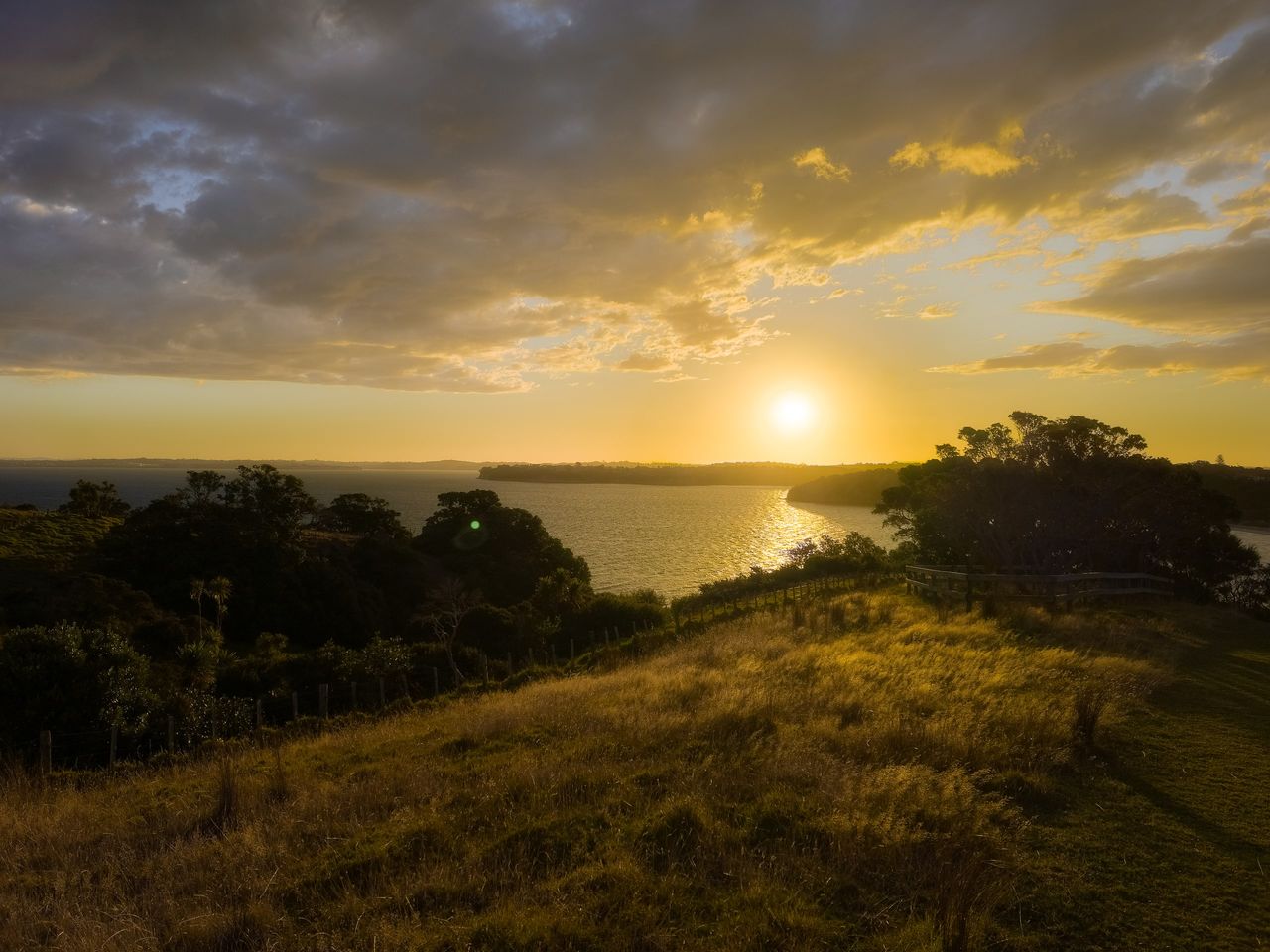Sun setting over a calm bay, seen from a grassy hilltop with a wooden fence, dark trees and golden clouds above