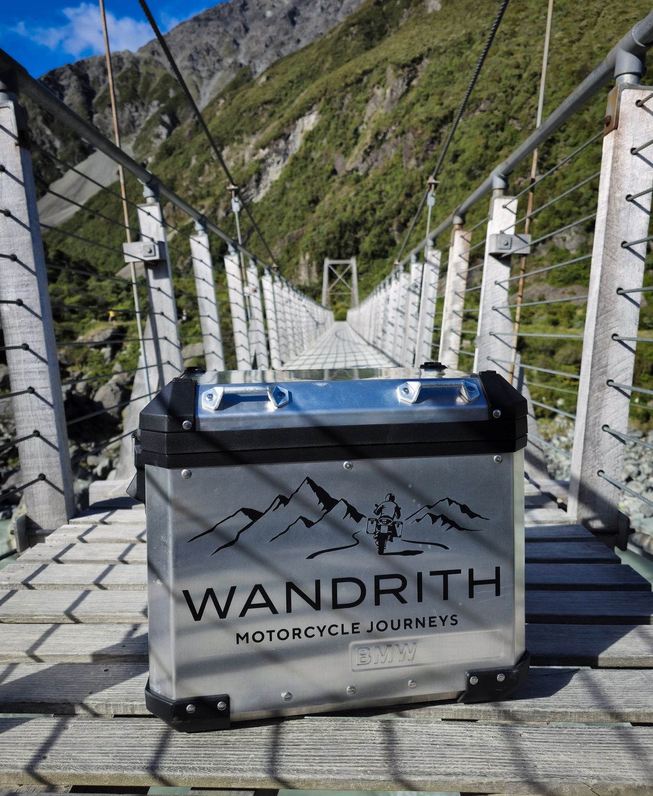 Wandrith-branded aluminium BMW pannier on the deck of a long swing bridge, with wire railings and a steep, bush-covered mountainside behind