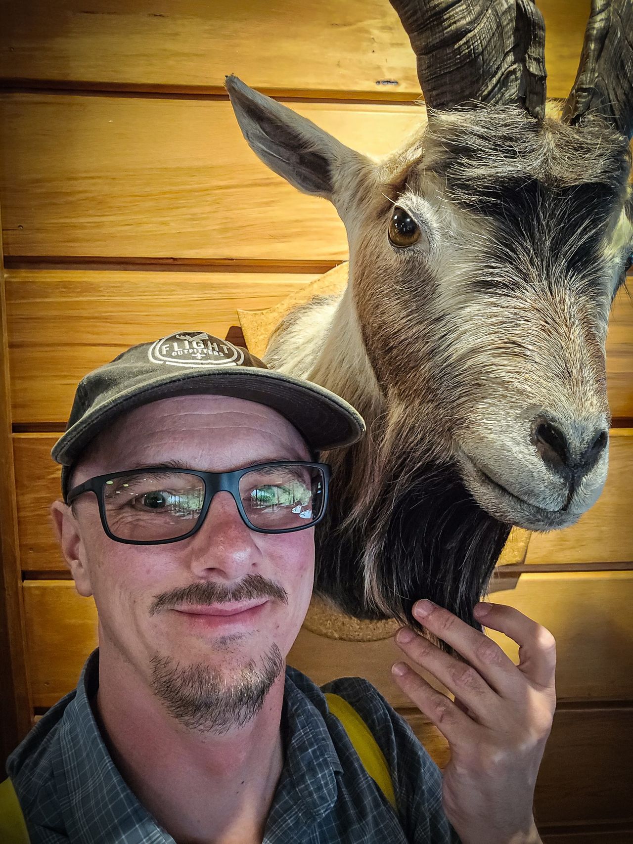 Smiling man in a cap and glasses taking a selfie beside a mounted goat head on a timber-panelled wall
