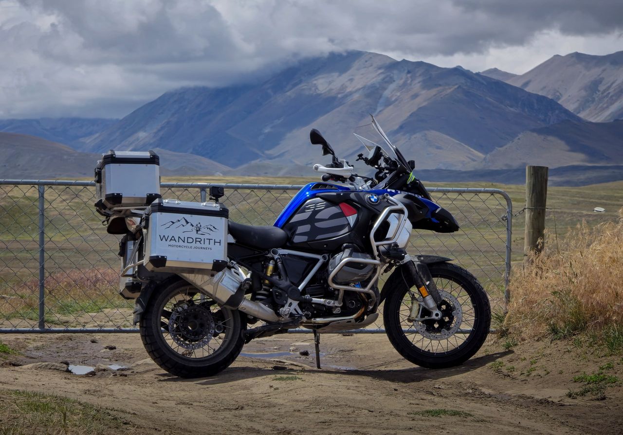Blue and black BMW R 1250 GS Adventure with Wandrith luggage parked by a farm gate, cloud-capped brown mountains behind