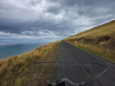 Rider’s view over a windscreen along a narrow road on a steep tussock hillside, high above a grey sea under heavy cloud