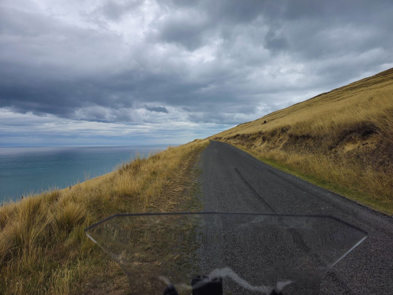 Rider’s view over a windscreen along a narrow road on a steep tussock hillside, high above a grey sea under heavy cloud
