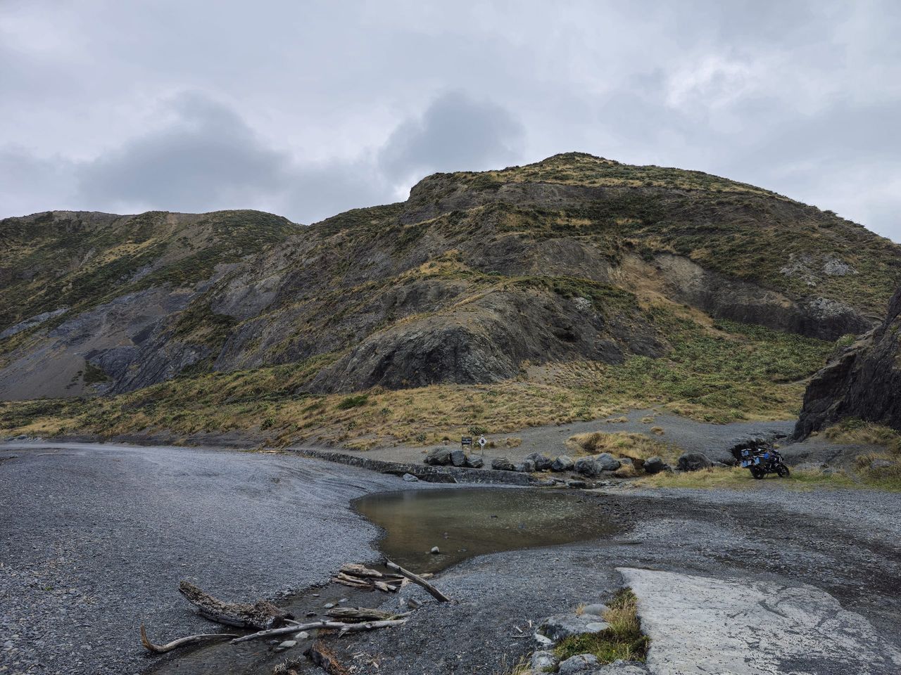 Loaded adventure motorcycle, small in frame, parked by boulders below steep scrubby hills, beside a grey gravel shore with driftwood